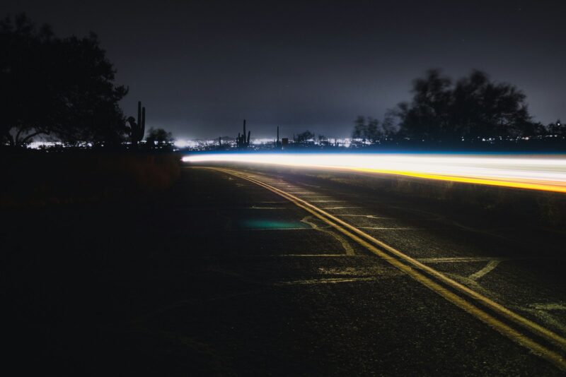 Enhanced Night Visibility on Roads – A Key Safety Feature for Hamilton Drivers Long exposure of a car's headlights and tail lights creating streaks of light along a dark, empty road at night, with city lights in the far distance.