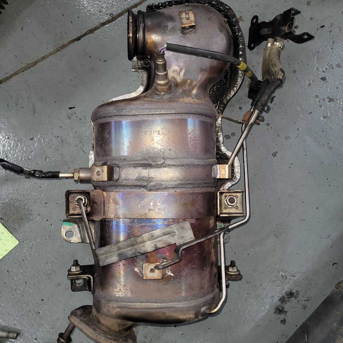A used catalytic converter with sensors and pipes attached lies on a workshop floor.