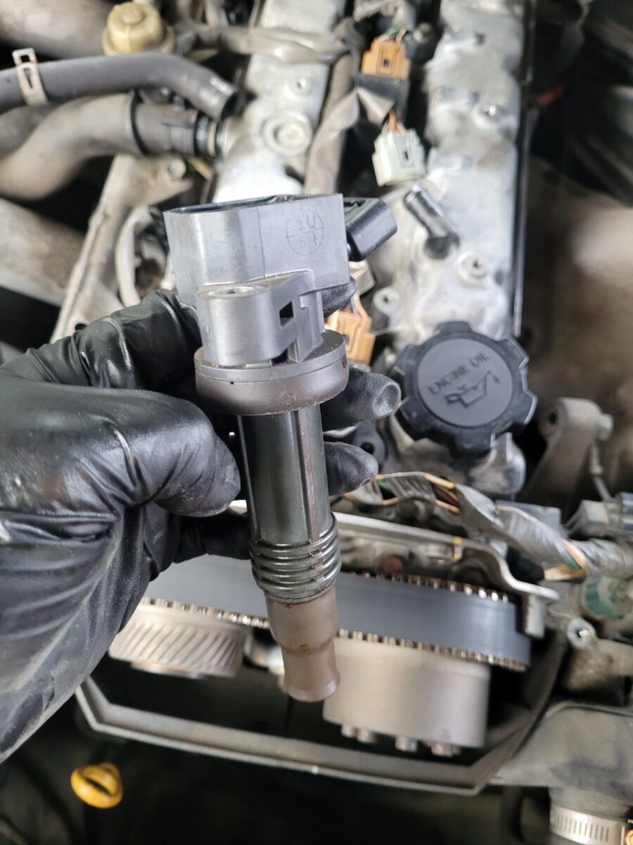 A gloved hand holds a car ignition coil over an engine.