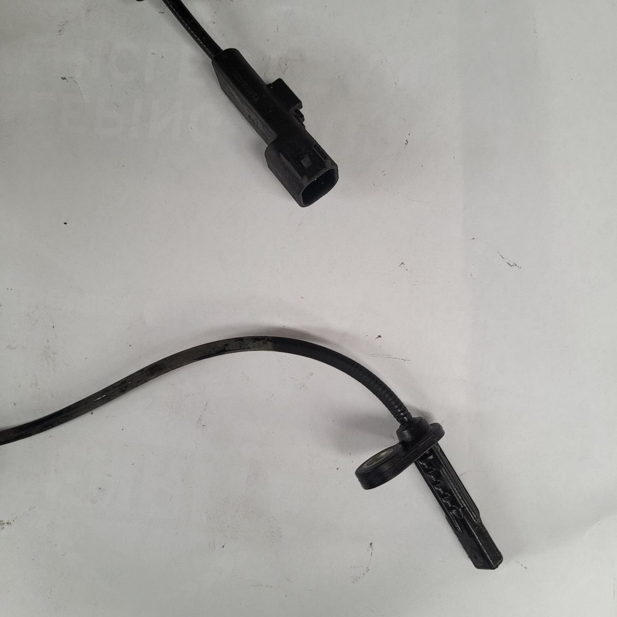 A black automotive ABS sensor with connector and cable, lying on a white surface.