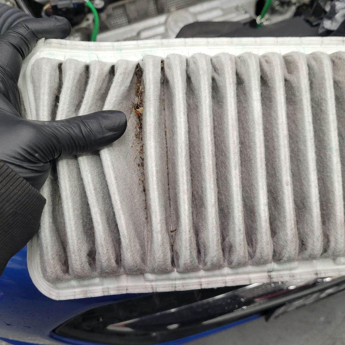 Hand wearing black glove holding a dirty pleated air filter with debris trapped inside.