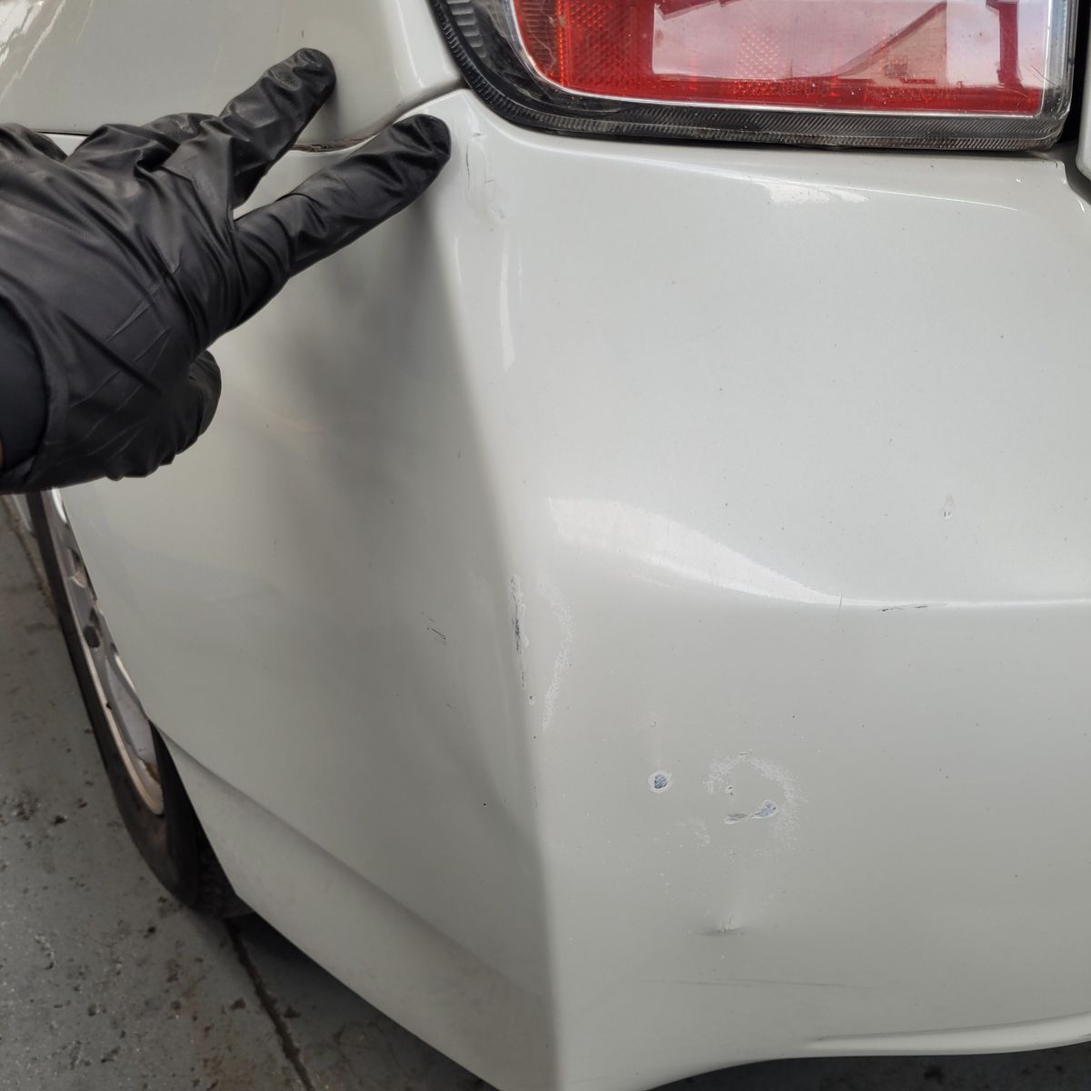 Gloved hand pointing at dent and scratches on white car bumper near taillight.