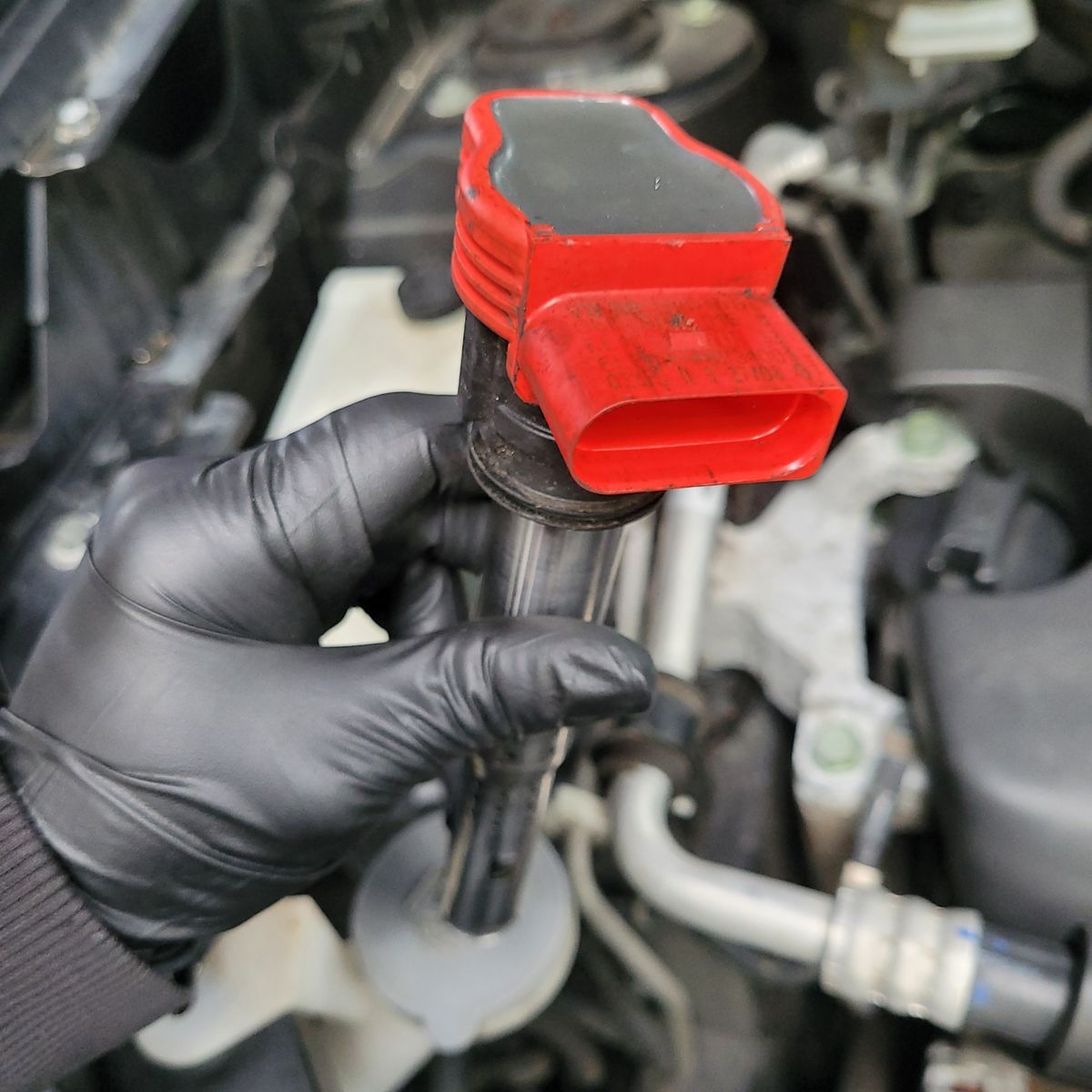 Mechanic Holding an Electronic Ignition Coil Gloved hand holding a red electronic ignition coil above an engine bay