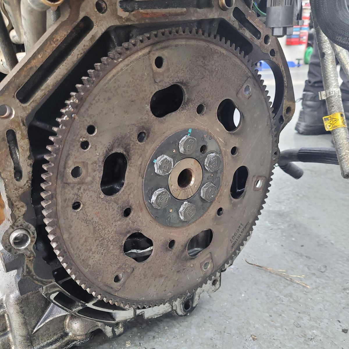 Rusty flywheel mounted on a car engine's crankshaft with bolts and teeth visible.
