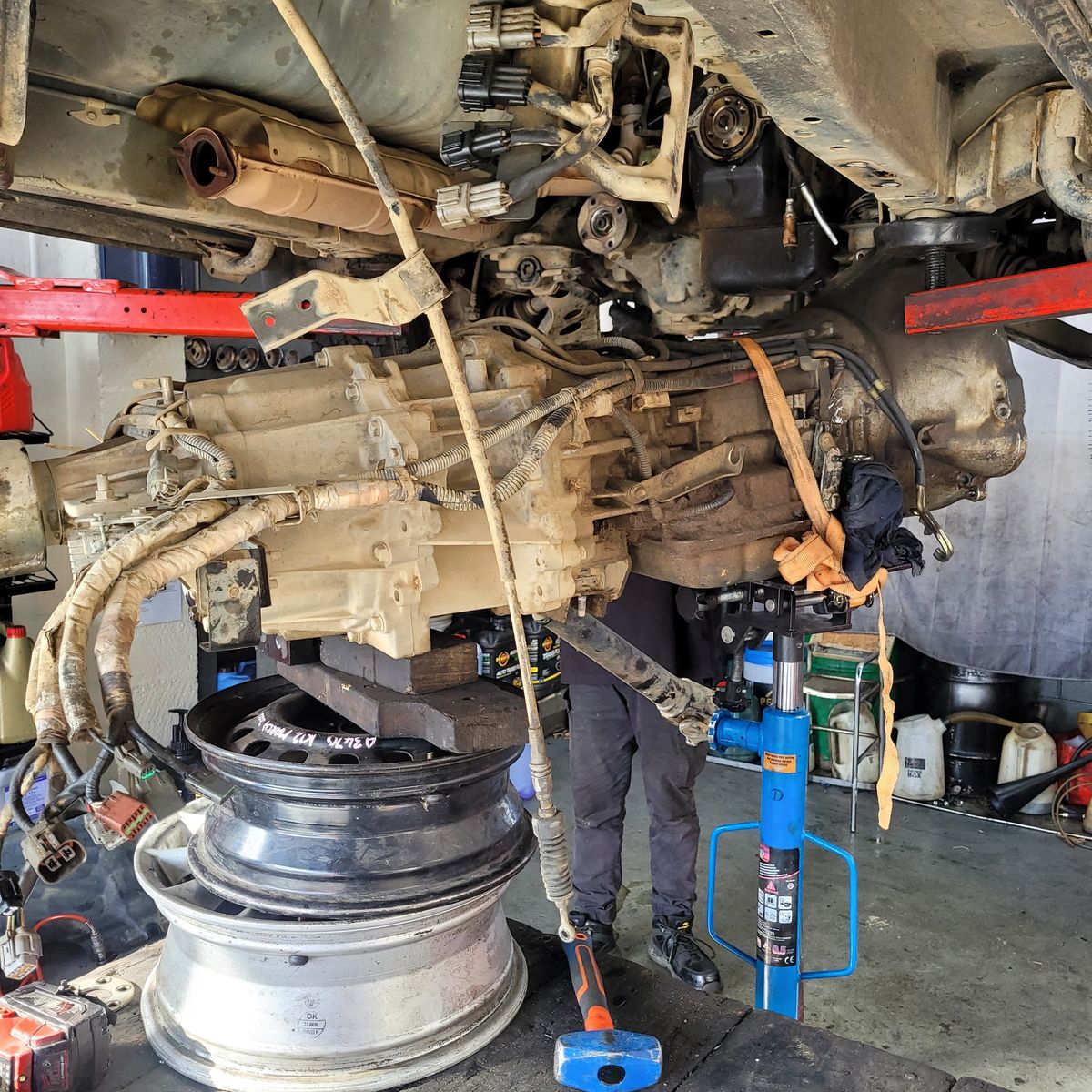 A car gearbox is being removed underneath a lifted vehicle using a jack and old wheel rims for support.