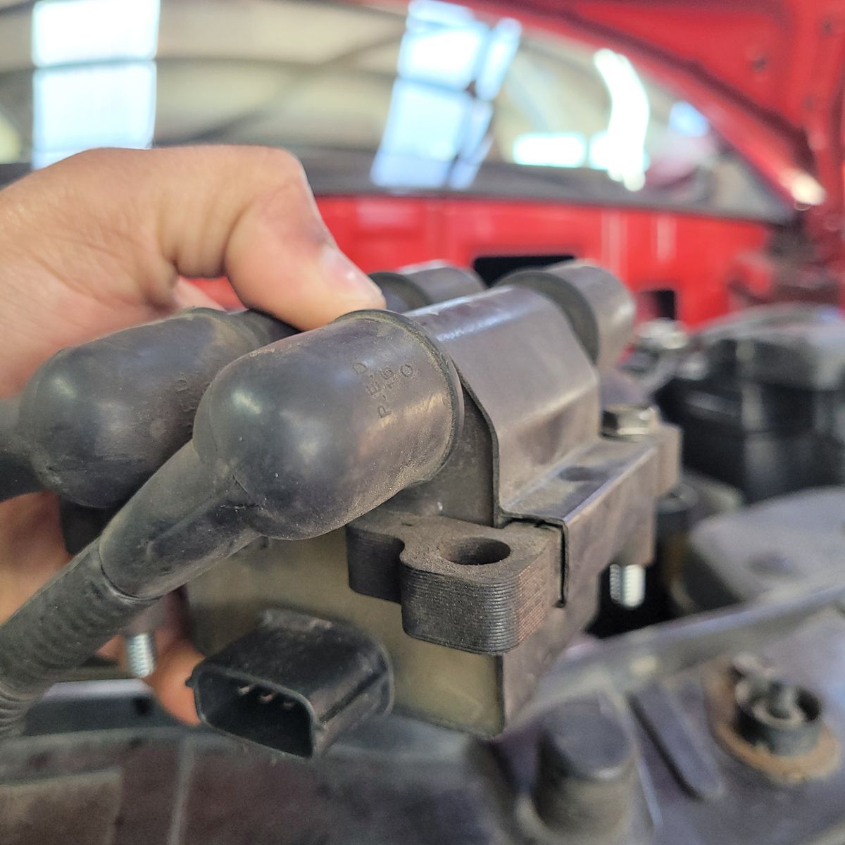 Hand holding a car ignition coil with attached spark plug wires inside an engine bay.