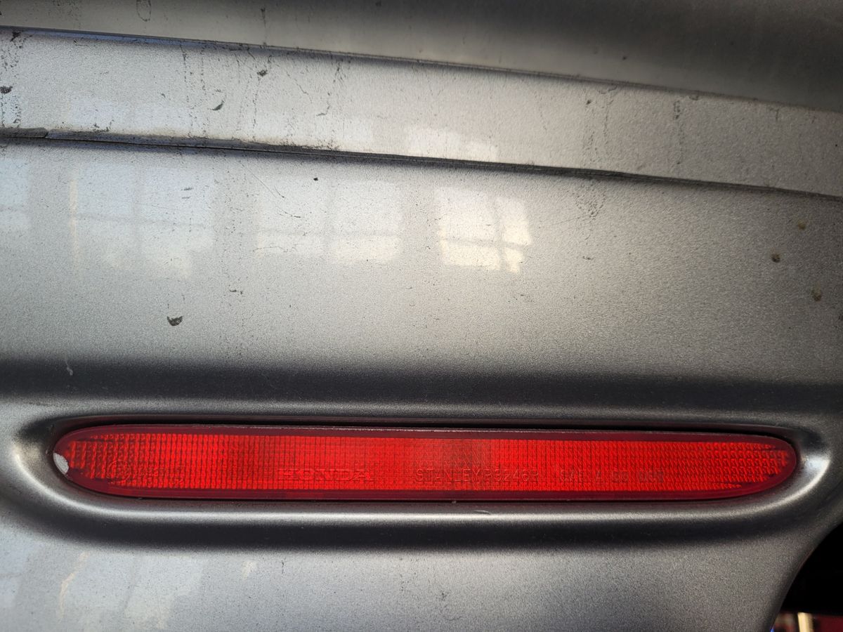 Close-up of a red reflector on a dusty silver car bumper.
