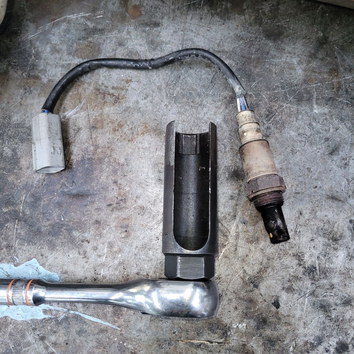A used oxygen sensor and socket wrench placed on a dirty metal surface.