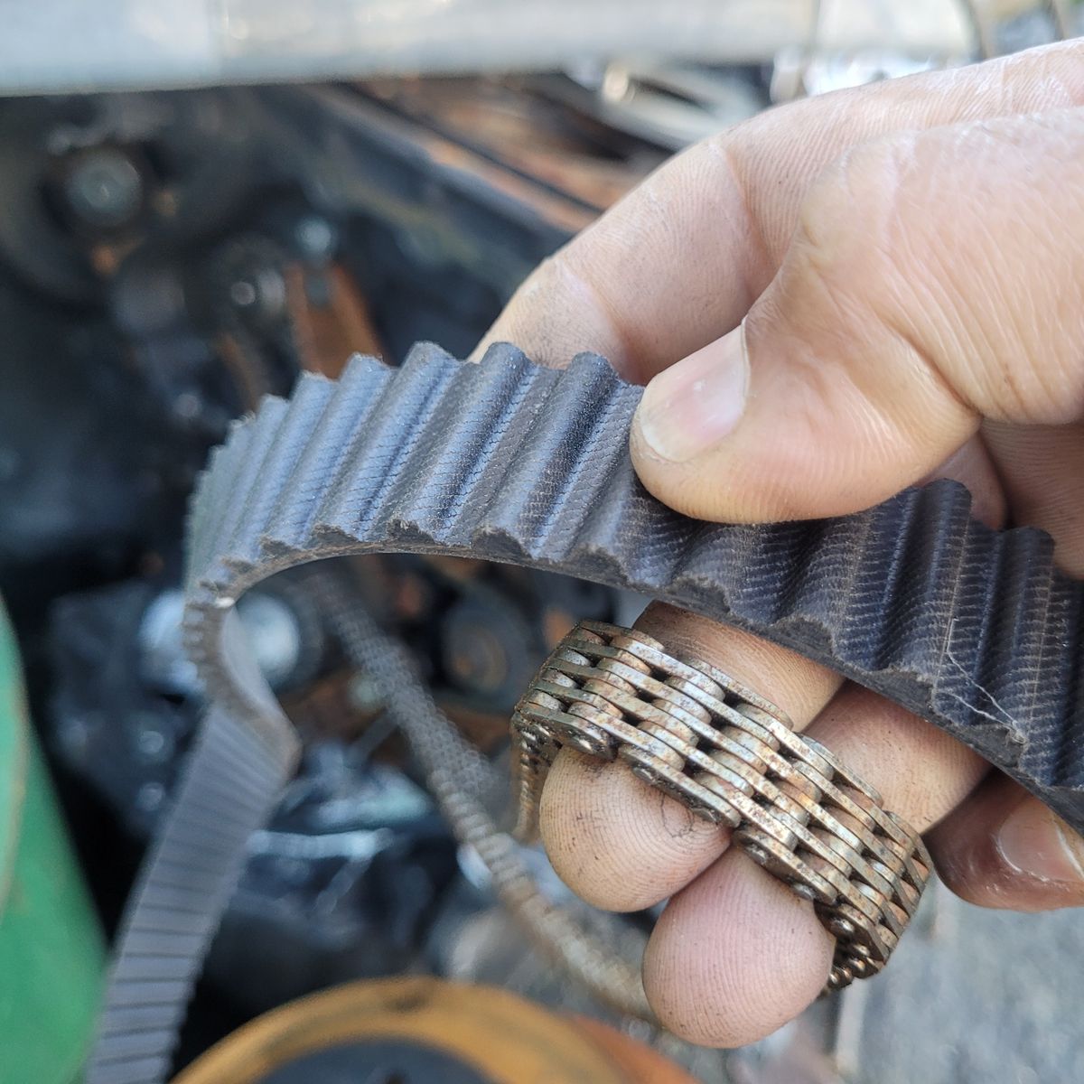 Comparing a Timing Belt and Timing Chain Hand holding a timing belt and timing chain in an engine bay