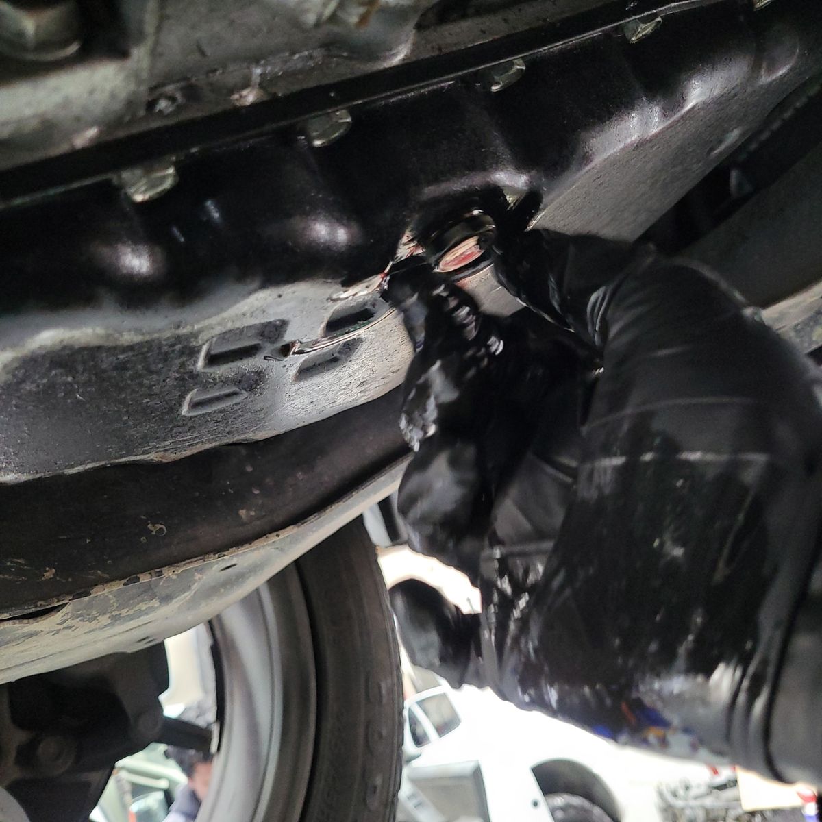 Mechanic's gloved hand checking transmission oil drain plug underneath a car